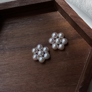 Könnte beinhalten: Ein Paar Perlenohrringe in Blumenform. Die Ohrringe bestehen aus kleinen, weißen Perlen, die in einem kreisförmigen Muster angeordnet sind. Die Ohrringe liegen auf einer Holzoberfläche.