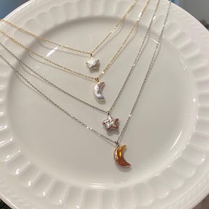Puede incluir: Cuatro delicados collares con colgantes celestiales, incluyendo estrellas y medias lunas. Las cadenas son doradas y plateadas, y los colgantes tienen un acabado nacarado. Los collares se exhiben en un plato blanco con borde festoneado.