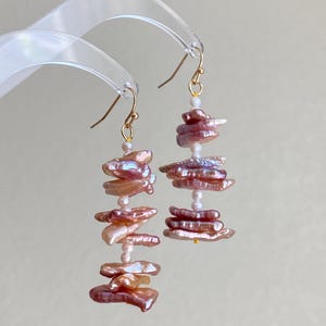 Puede incluir: Un par de pendientes colgantes con perlas apiladas, de forma irregular, de color marrón rosado. Los pendientes tienen ganchos dorados y pequeñas cuentas blancas entre las pilas de perlas. El fondo es de un color suave y neutro.