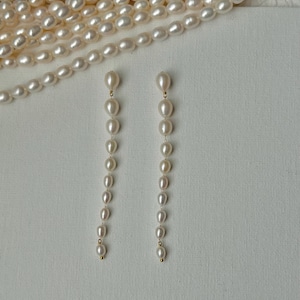 May include: Elegant pearl drop earrings with a gold accent. The earrings feature a series of graduated, oval-shaped pearls suspended from a delicate gold post. Several strands of pearls are visible in the background, suggesting a jewelry collection.