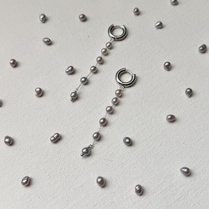 May include: A pair of silver hoop earrings with a dangling chain of gray pearls. The earrings are displayed on a white surface scattered with loose gray pearls, creating a minimalist aesthetic. The earrings are a delicate piece of jewelry.