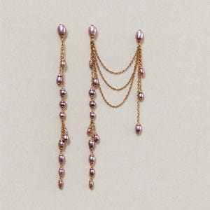 May include: A pair of gold-toned chain earrings featuring dangling lavender-colored oval beads. One earring consists of a single vertical strand of beads, while the other features three draped chain segments connecting two vertical beaded strands.