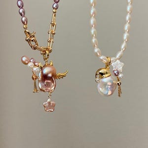 May include: Two pearl necklaces. One features a gold chain with a pink baroque pearl pendant with wings and a star. The other necklace has a white pearl strand with a gold pendant and a large white pearl.
