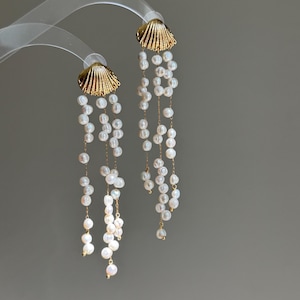Può includere: Un paio di lunghi orecchini pendenti con perni in metallo dorato a forma di conchiglia. Ogni orecchino presenta diverse delicate catenelle dorate che scendono dalla conchiglia, decorate con piccole perle d'acqua dolce bianche e irregolari.