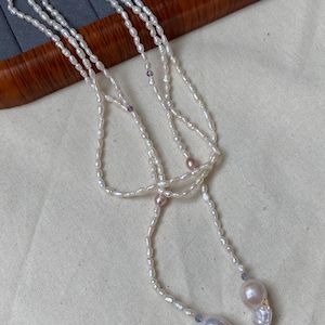 May include: A delicate white pearl necklace with a lariat design. The necklace features small, oval-shaped pearls strung together, with larger, baroque pearls at the ends. Accents include pink and purple beads.