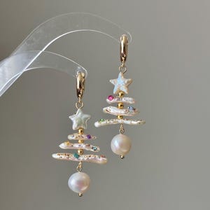 May include: A pair of gold hoop earrings featuring dangling charms shaped like Christmas trees. Each tree is constructed from white baroque pearl segments, small gold beads, and tiny colorful gemstone accents, topped with a star-shaped pearl.