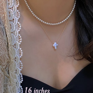 May include: A layered necklace featuring two strands of pearls and a gold chain with a cross pendant. The necklace is approximately 16 inches long. The image shows the necklace on a person's neck.