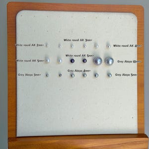 May include: A selection of Akoya pearls, displayed on a cream-coloured surface. The pearls vary in size from 2mm to 10mm, with white and grey hues. Text labels indicate the size and type of each pearl.