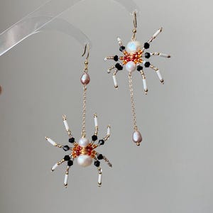 May include: A pair of spider-shaped earrings. Each earring features a pearl body, black and white beaded legs, and a gold-colored wire frame. One earring has a dangling pink pearl. The earrings are designed with a gold-colored hook.