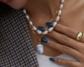 May include: A layered necklace featuring white baroque pearls and a gold chain with a large, rectangular pearl pendant attached by a toggle clasp. A gold ring with a central white stone is worn on the finger. The jewelry is worn over a white top.