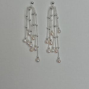 Op de afbeelding: Elegante zilveren oorbellen met trapsgewijze strengen van kleine zilveren kralen en glanzende witte parels. De oorbellen hebben een delicaat ontwerp, met meerdere strengen parels en kralen die aan een kleine zilveren bal aan de bovenkant hangen.