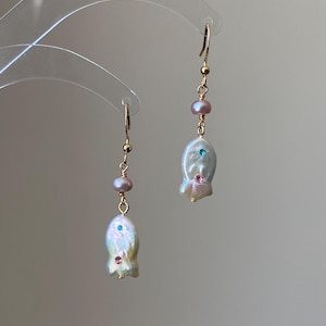 Op de afbeelding: Een paar goudkleurige oorhangers. Elke oorbel heeft een grote, onregelmatige witte parel met een kleine blauwe en roze edelsteen. Boven de parel zitten twee kleinere parels en een goudkleurige haak.