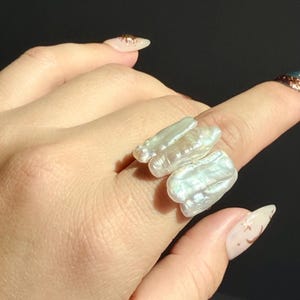 Könnte beinhalten: Ein Ring mit drei länglichen, unregelmäßigen weißen Süßwasserperlen mit einem stark glänzenden, irisierenden Finish. Die Perlen sind nebeneinander angeordnet und erzeugen ein strukturiertes, organisches Erscheinungsbild mit sichtbaren Perlmuttschichten.