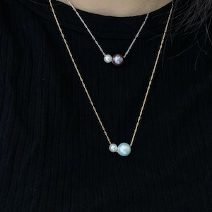 May include: Two delicate necklaces with pearl pendants. One necklace features a gold chain with a white and a light blue pearl. The other necklace has a silver chain with a white and a dark purple pearl. The necklaces are displayed against a black background.