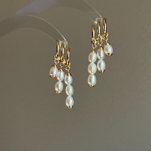 May include: A pair of gold hoop earrings, each featuring three dangling charms made of white oval freshwater pearls. The pearls are attached to the gold hoops with small gold wire wraps, creating a tiered, cascading effect of varying lengths.