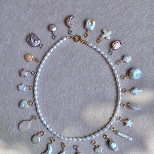 Op de afbeelding: Een parelketting met een gouden sluiting, omringd door diverse parelbedels. De bedels omvatten sterren, manen, harten en abstracte vormen, allemaal in tinten wit, crème en iriserend. De ketting en bedels liggen op een lichtbeige oppervlak.
