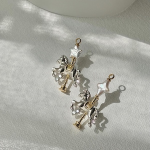 Puede incluir: Un par de pendientes de caballo de carrusel plateados con detalles dorados y una parte superior en forma de perla. Los pendientes presentan un diseño detallado de un caballo de carrusel con una perla en forma de estrella en la parte superior. Sobre fondo blanco.