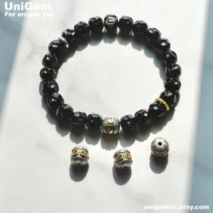 May include: Black beaded bracelet with gold accents. The bracelet features a unique design with a silver charm and a gold ring. The bracelet is shown with two extra charms.
