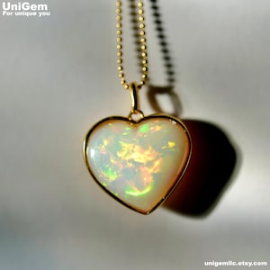 May include: A heart-shaped opal pendant set in a gold bezel, suspended from a matching gold ball chain. The opal displays a milky white base with vibrant flashes of green, yellow, and orange iridescence throughout the stone's smooth surface.