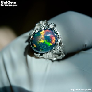May include: A silver ring featuring a large, oval-shaped black opal with vibrant flashes of red, green, and yellow fire. The metal band has a textured, organic design resembling small branches or twigs surrounding the central gemstone.