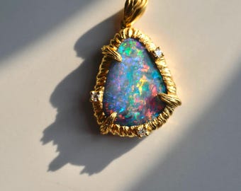 May include: A gold pendant featuring a pear-shaped opal with vibrant blue, green, and red flashes. The stone is set in a textured gold frame with four prongs, each accented by a small clear gemstone. A gold bail is attached to the top of the pendant.