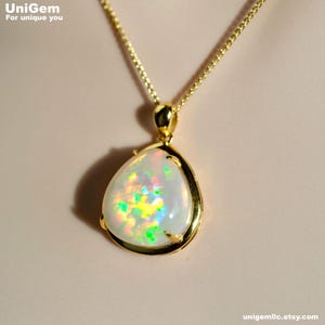 May include: A teardrop-shaped opal pendant set in a polished yellow gold frame, suspended from a matching yellow gold chain. The opal displays a vibrant play-of-color with flashes of green, yellow, and orange against a milky white base.
