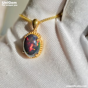 May include: A gold pendant necklace featuring an oval black opal gemstone with red and green flashes. The opal is set in a gold frame with a textured border. The chain is also gold. The Unigem logo is in the top left corner.