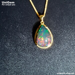 May include: A teardrop-shaped pendant necklace featuring a dark, iridescent opal gemstone with flashes of green, red, and orange color play. The stone is set in a gold-toned metal frame with a beaded edge, suspended from a matching gold-toned chain.