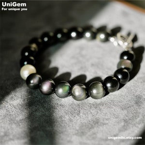 May include: A black and iridescent gemstone bracelet with a silver clasp. The bracelet has a single white pearl bead.