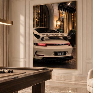 May include: A large rectangular wall art print featuring the rear view of a white Porsche sports car with a prominent rear wing, red taillights, and a license plate reading 412, displayed in a room with a pool table and a cream-colored armchair.