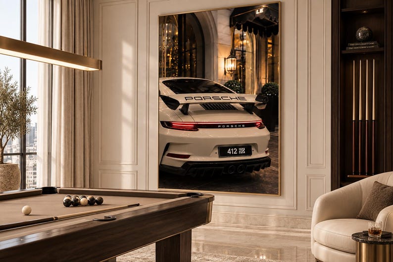 May include: A large rectangular wall art print featuring the rear view of a white Porsche sports car with a prominent rear wing, red taillights, and a license plate reading 412, displayed in a room with a pool table and a cream-colored armchair.