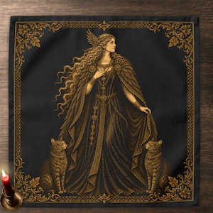 May include: A square black fabric cloth featuring a gold-toned illustration of a long-haired figure wearing a winged headpiece and a flowing gown, flanked by two cats. The design is enclosed within an ornate gold-toned border of leaves and knots.