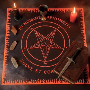 May include: A black altar cloth with an orange border and a pentagram design. The text "IN NOMINE APHOMETIS SOLVE ET COA" is printed in orange. A lit black candle, a red candle, a chalice, crystals, a book, and a dagger are on the cloth.