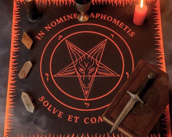 May include: A black altar cloth with an orange border and a pentagram design. The text "IN NOMINE APHOMETIS SOLVE ET COA" is printed in orange. A lit black candle, a red candle, a chalice, crystals, a book, and a dagger are on the cloth.