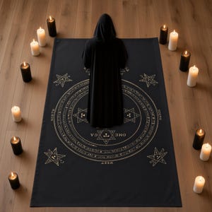 May include: A black rectangular mat featuring a gold circular design with a central hexagram, the word OMEGA, and four smaller stars at the corners. A figure in a black hooded robe stands on the mat, surrounded by white and black pillar candles.