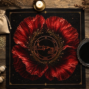 May include: A square black fabric cloth featuring a large, vibrant red poppy flower design. In the center, a circular emblem displays a pointing hand, Hebrew text, and a star symbol, all surrounded by a delicate golden branch wreath.