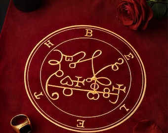 May include: A burgundy cloth with a gold embroidered sigil, a black candle holder, a red rose, and a gold ring with a black stone. The sigil is circular with stylized lettering and symbols.