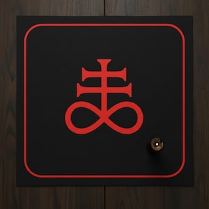 May include: Black square sign featuring a red border and a red symbol. A small, gold-coloured knob is on the right side. The sign is mounted on a dark wood surface.