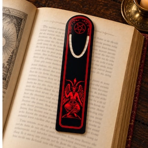 May include: A black bookmark with a rounded top and a U-shaped cutout. The design features a red pentagram at the top and a red illustration of a winged, horned figure sitting in a cross-legged position in the center, framed by red lines.