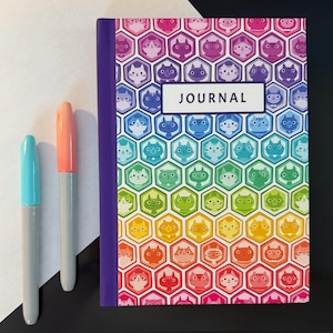 May include: A colorful journal with a cat-themed design, featuring a rainbow gradient of hexagonal cells, each containing a cat face. The journal has a purple spine and the word "JOURNAL" on the cover. Two markers, one blue and one orange, are next to the journal.