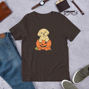 Könnte beinhalten: Braunes T-Shirt mit einer Cartoon-Illustration eines gelben Labrador-Welpen, der in einem geschnitzten orangefarbenen Kürbis sitzt. Der Kürbis hat ein Kürbisgesicht und ist von kleineren Kürbissen und grünen Blättern umgeben. Festliches Design.