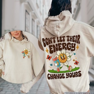 May include: Sand-colored hoodie with a front pocket and drawstring hood. The back features a retro graphic of a smiling flower holding a battery, with the text "DON'T LET THEIR ENERGY CHANGE YOURS". A smaller version of the graphic is on the front.