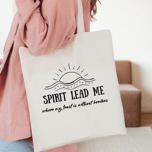 May include: White tote bag with black text that reads "Spirit Lead Me where my trust is without borders" and a graphic of a sun rising over a wavy line.