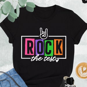 May include: Black t-shirt with a white graphic that says "ROCK the test" in colorful letters. The graphic also includes a hand making the rock and roll hand gesture.