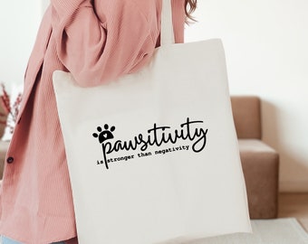May include: A white tote bag with black text that reads "Pawsitivity is stronger than negativity". The bag has a paw print design and is being held by a person wearing a pink sweater.