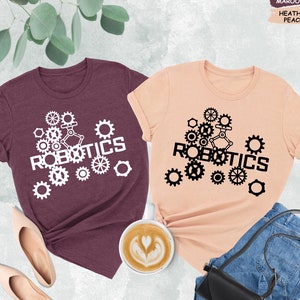 May include: Two t-shirts with the word "ROBOTICS" printed on them. The shirts are in heather maroon and heather peach colors. The word "ROBOTICS" is surrounded by gears.