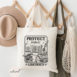 May include: A white tote bag with black text that reads "Protect Public Libraries". The bag has a graphic of a stack of books, a quill pen, and a magnifying glass. The bag is hanging on a wooden rack with a brown hat and two string bags.