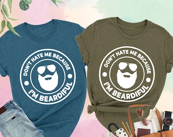 May include: Two t-shirts with a white graphic design. The design features a bearded man wearing sunglasses and the text "Don't hate me because I'm beardful". One shirt is teal and the other is olive green.