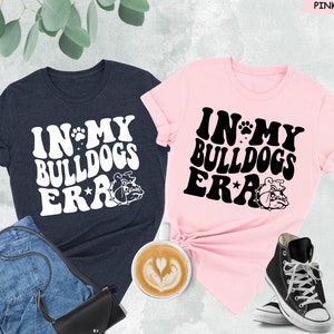 May include: Two t-shirts, one navy blue and one pink, with the text "In My Bulldogs Era" in a retro font. Each shirt features a cartoon bulldog graphic.