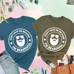 May include: Two t-shirts with a white graphic design. The design features a bearded man wearing sunglasses and the text "Don't hate me because I'm beardful". One shirt is teal and the other is olive green.